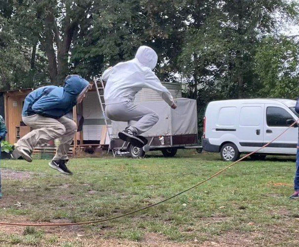 Zwei Personen springen draußen über ein Seil, mit Anhänger und Lieferwagen im Hintergrund.