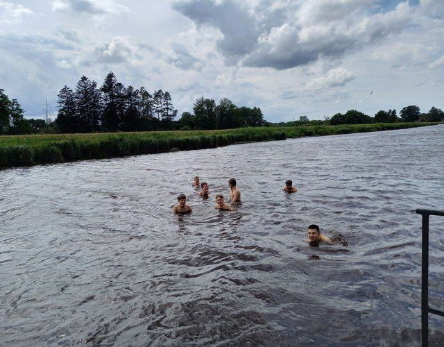 Jugendliche schwimmen in einem Fluss, mit grünen Ufern und bewölktem Himmel.